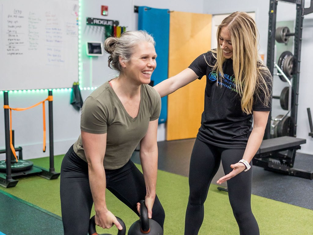 Personal trainer coaching a smiling client through a kettlebell deadlift to keep her muscle while losing weight